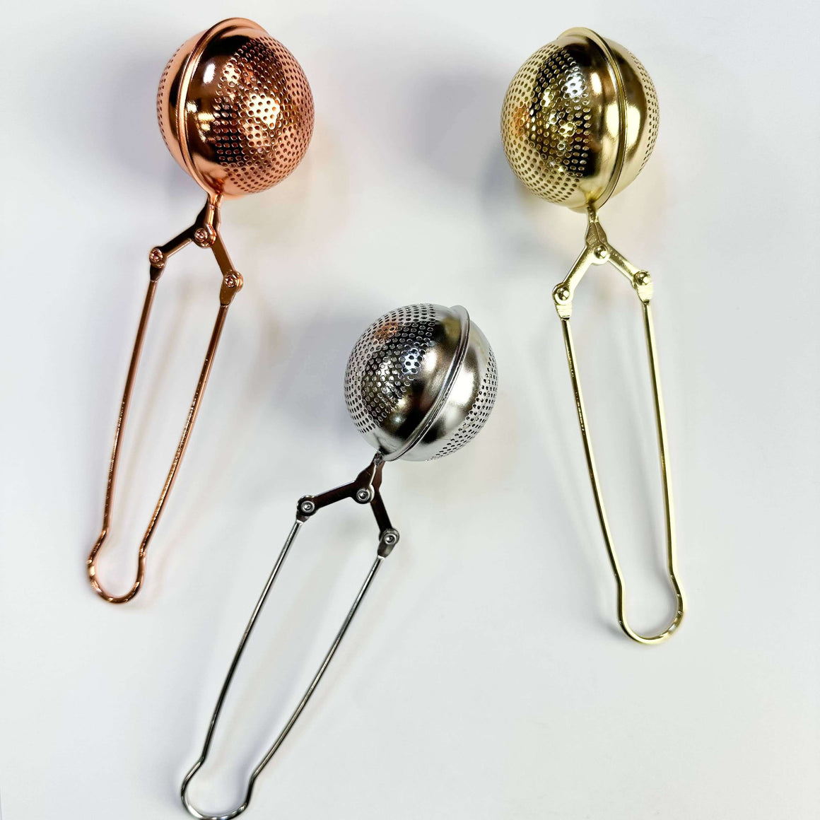 Three metal tea infusers in rose gold, silver, and gold on a white background
