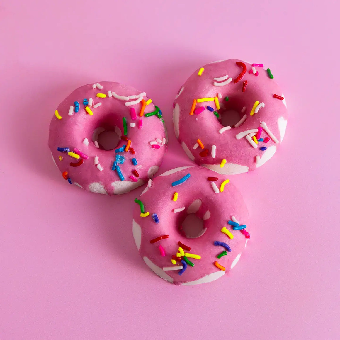 Birthday cake bath bombs shaped like colorful donuts with sprinkles on a pink background.