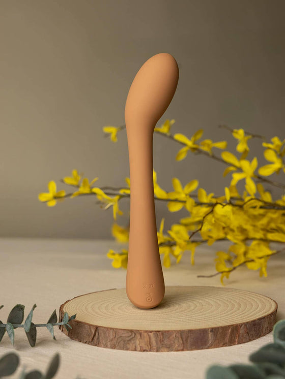 Cecily Curved G-Spot and Wand-Style Vibrator, a sleek curved g-spot vibrator on a natural wood stand surrounded by flowers.