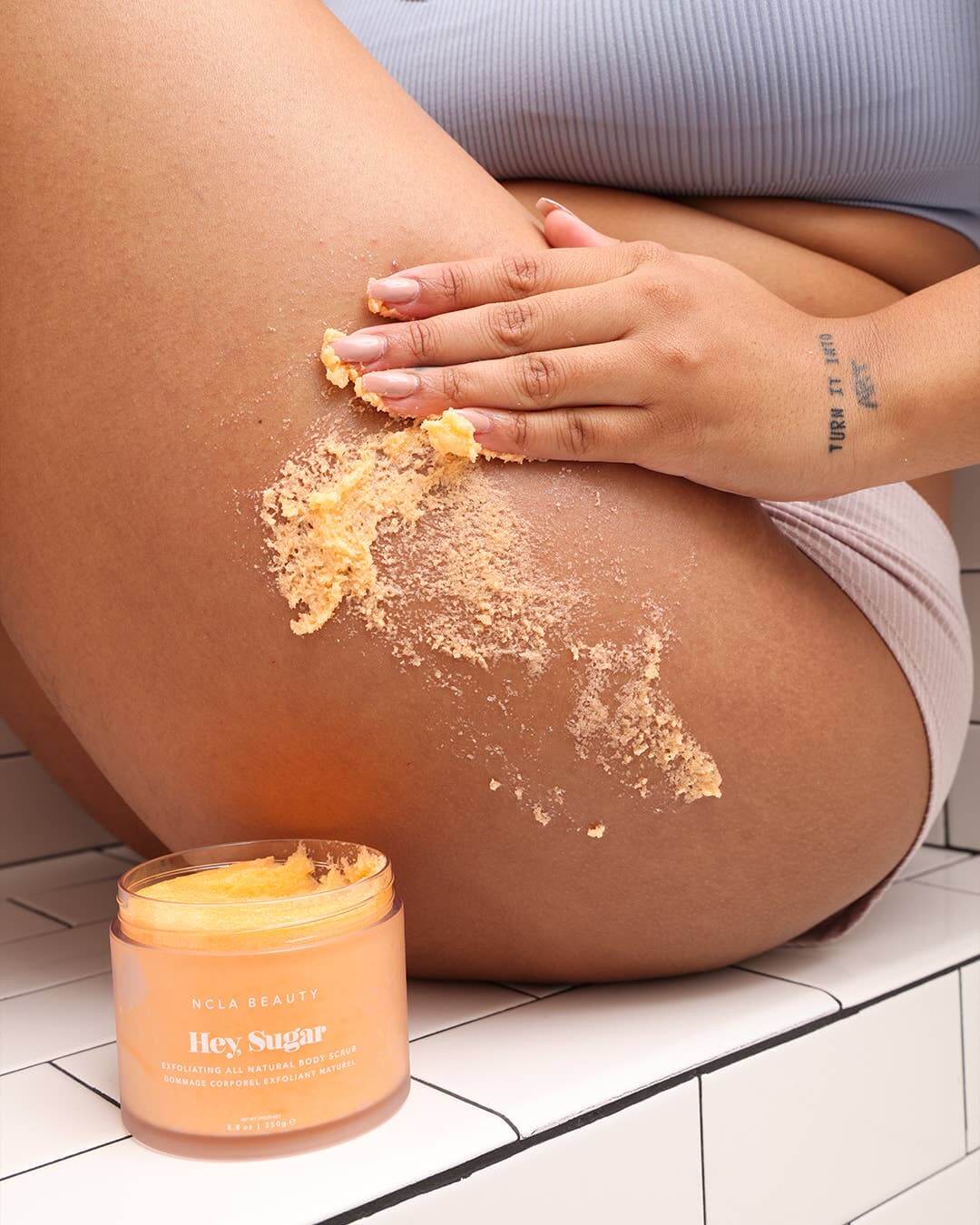 Person applying body scrub to their leg with a jar of the product next to them.