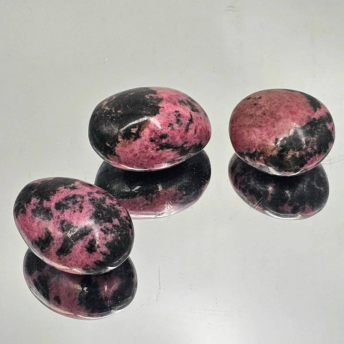 Three rhodonite stones on a reflective surface with a white background