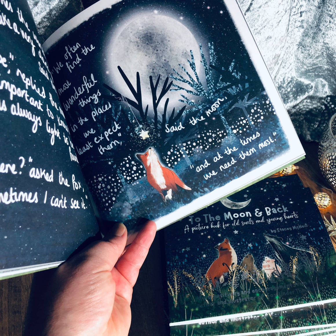 Open book with moon and fox illustration, held by a hand, next to closed book and candle on wooden surface.