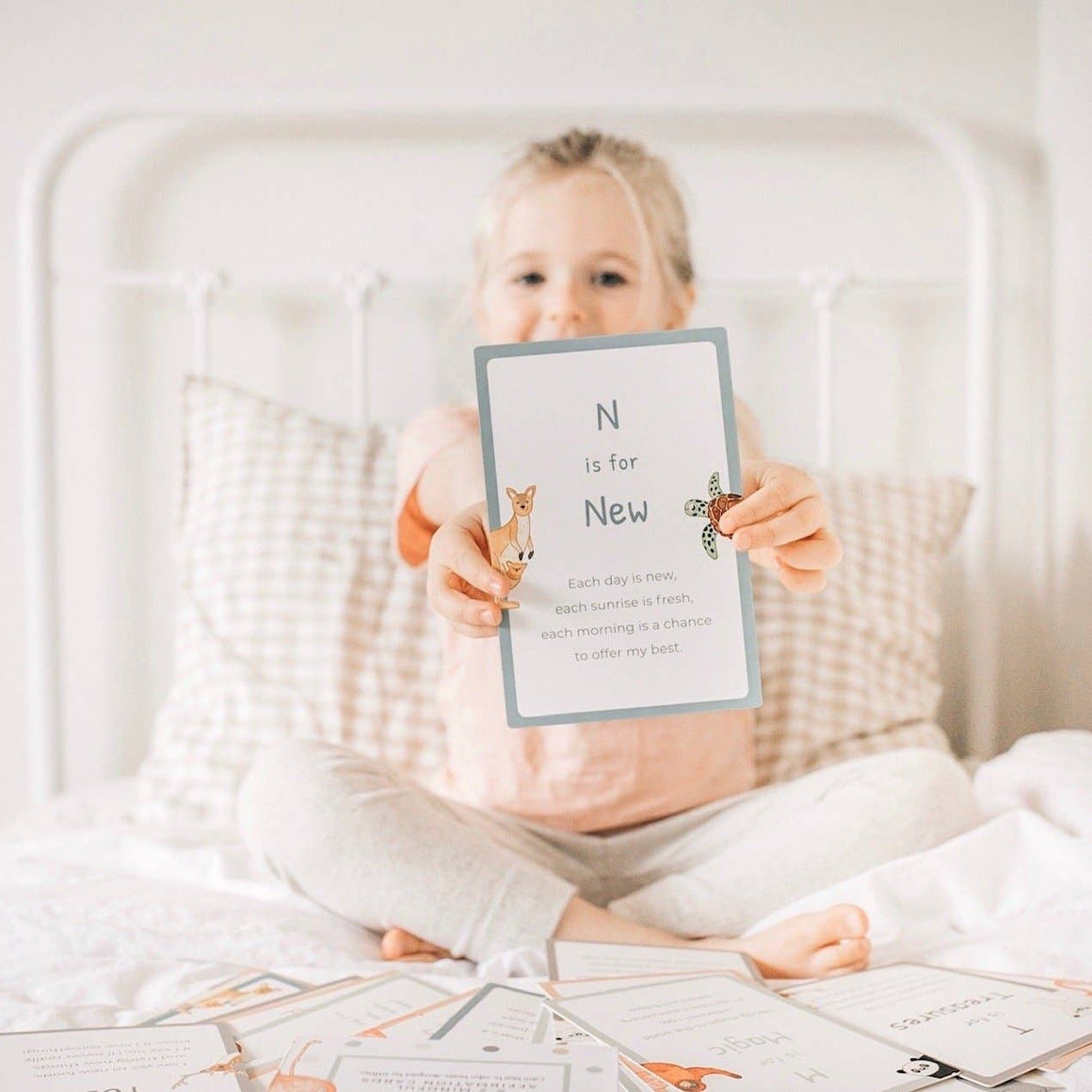 Child holding a card with 'N is for New' text on a bed