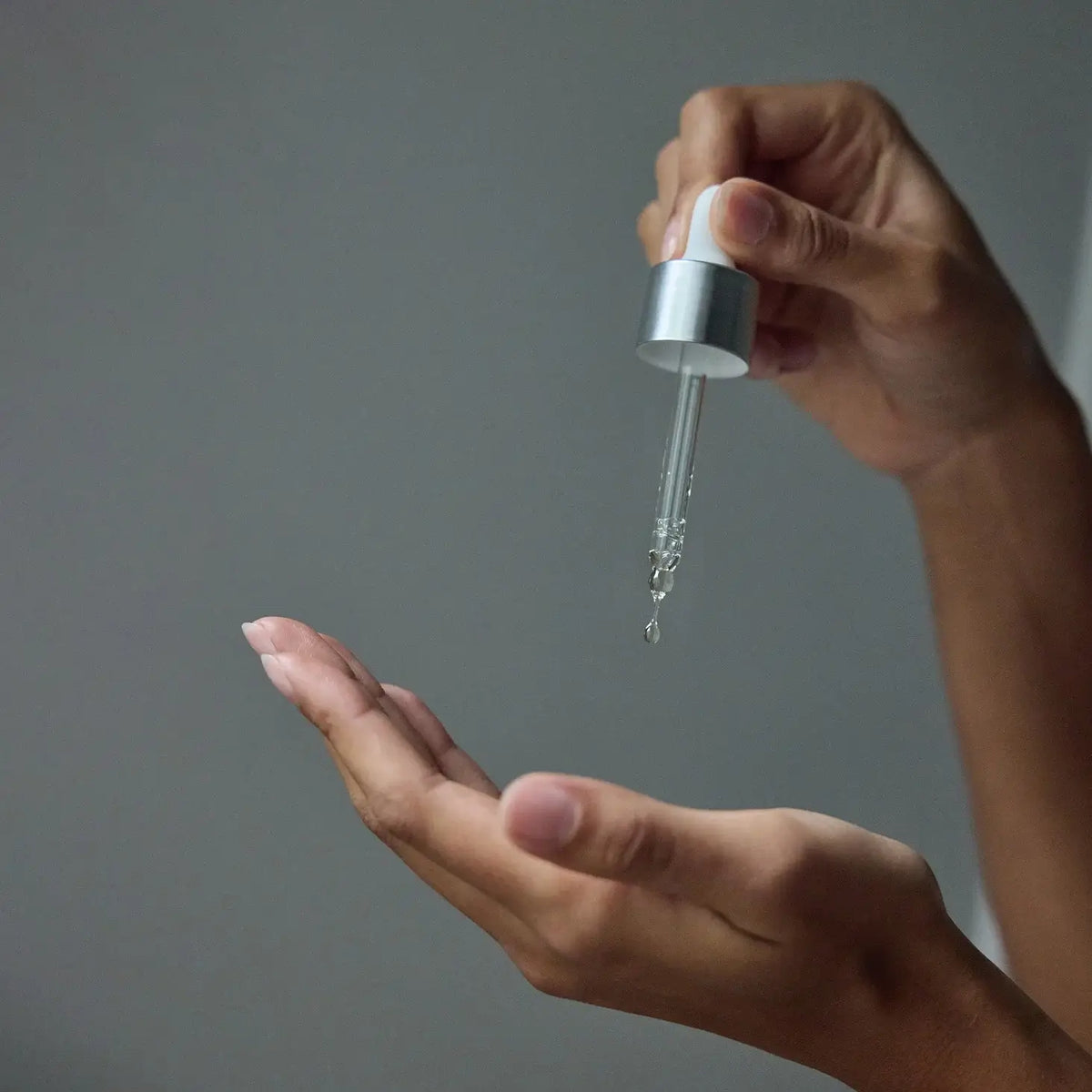 Hand holding a dropper with clear liquid over another hand against a gray background