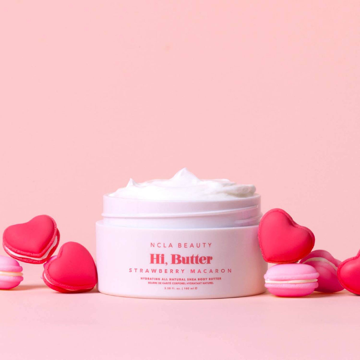 Jar of NCLA Beauty Hi, Butter Strawberry Macaron body butter with heart-shaped macarons on a pink background