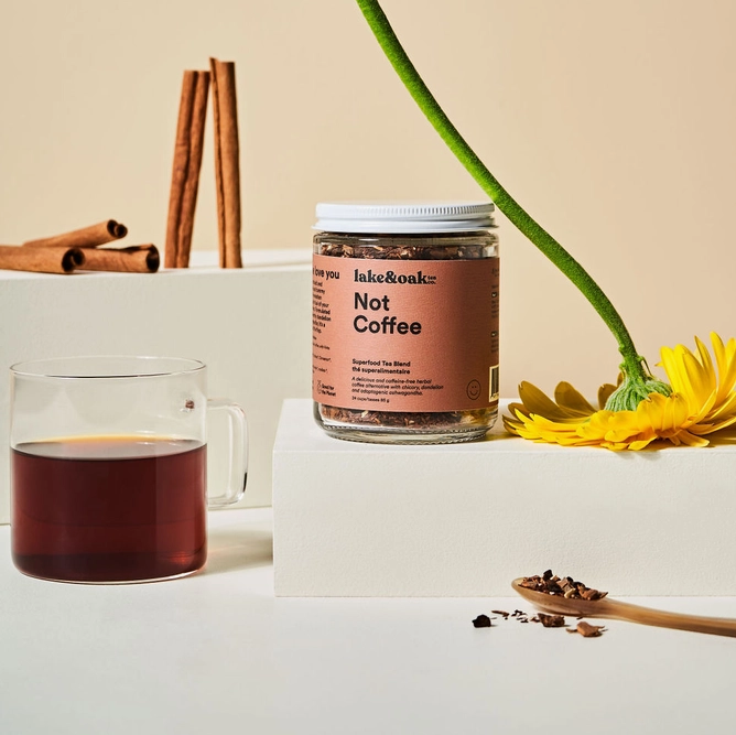 Jar of 'Not Coffee' product on a white surface with a glass of dark liquid, cinnamon sticks, and a yellow flower.