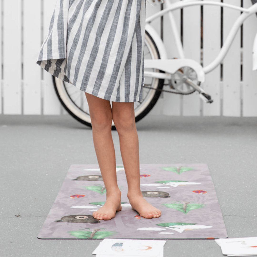 Nature Print Kids Yoga Mat on the floor with a child standing barefoot, surrounded by nature illustrations and a bicycle in the background.