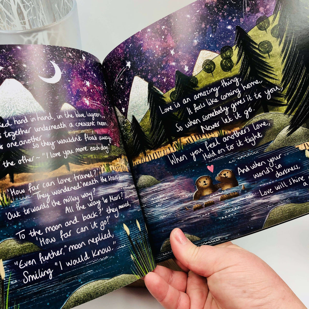 Open book with illustrated pages and a hand holding it, featuring a night sky and mountain landscape.