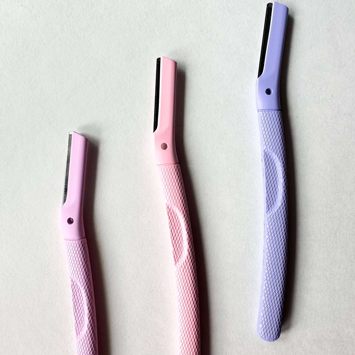 Three eyebrow razors in pink and purple on a light gray background