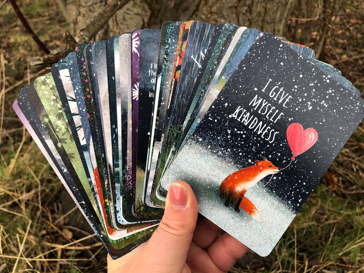 A hand holding a fan of colorful Positive Affirmation Cards featuring inspiring messages and artwork.