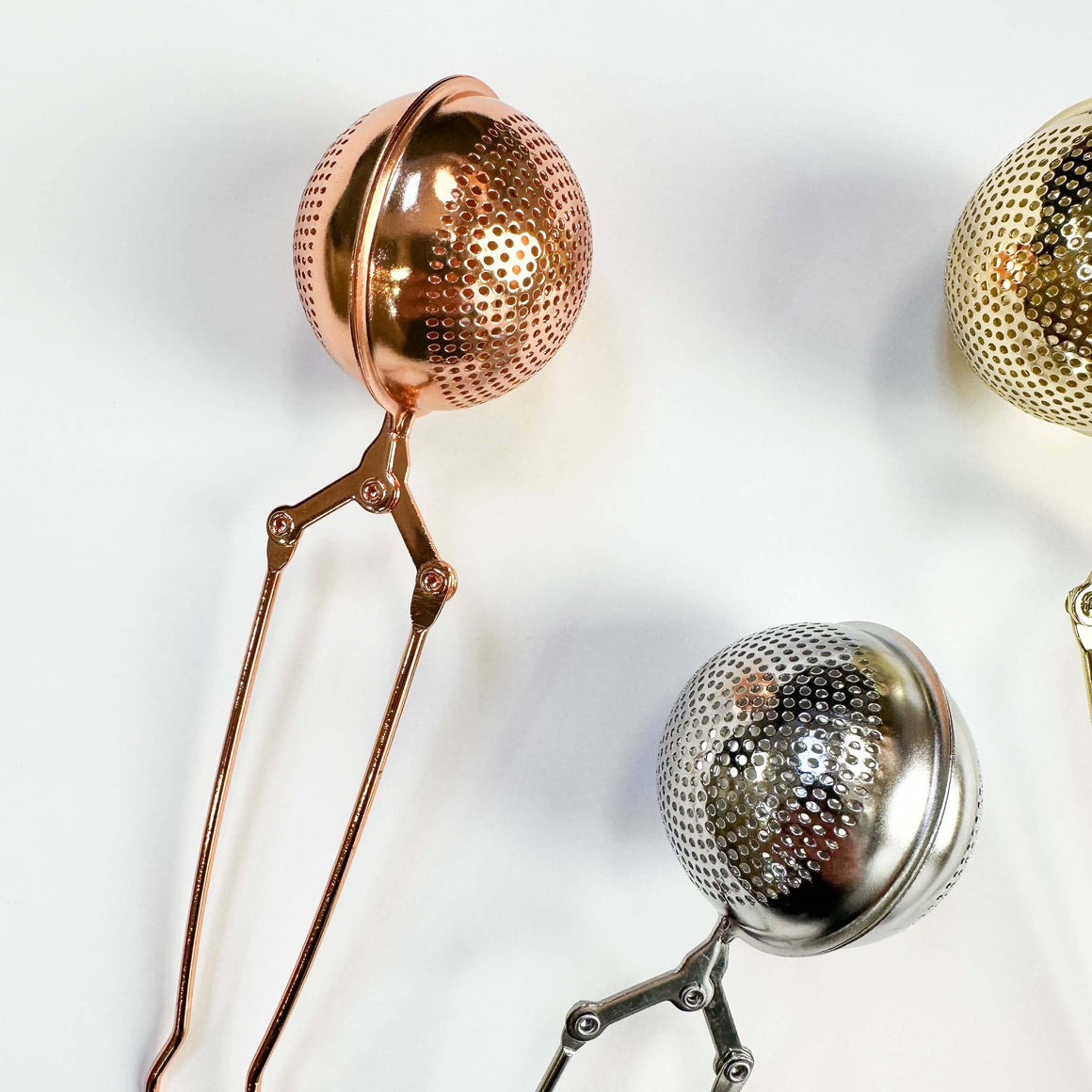 Metallic tea infusers in rose gold, silver, and gold on a light background