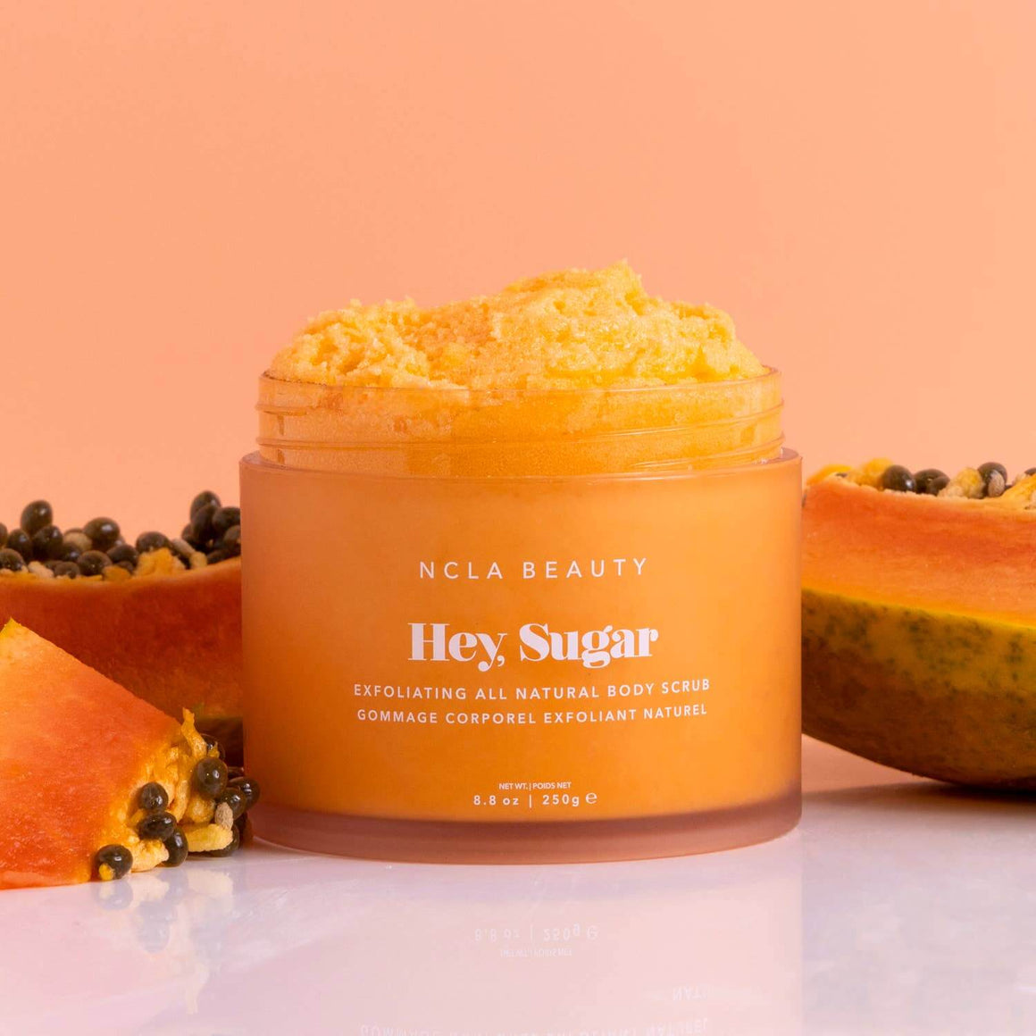 Orange jar of NCLA Beauty 'Hey Sugar' body scrub with papaya and seeds on a peach background
