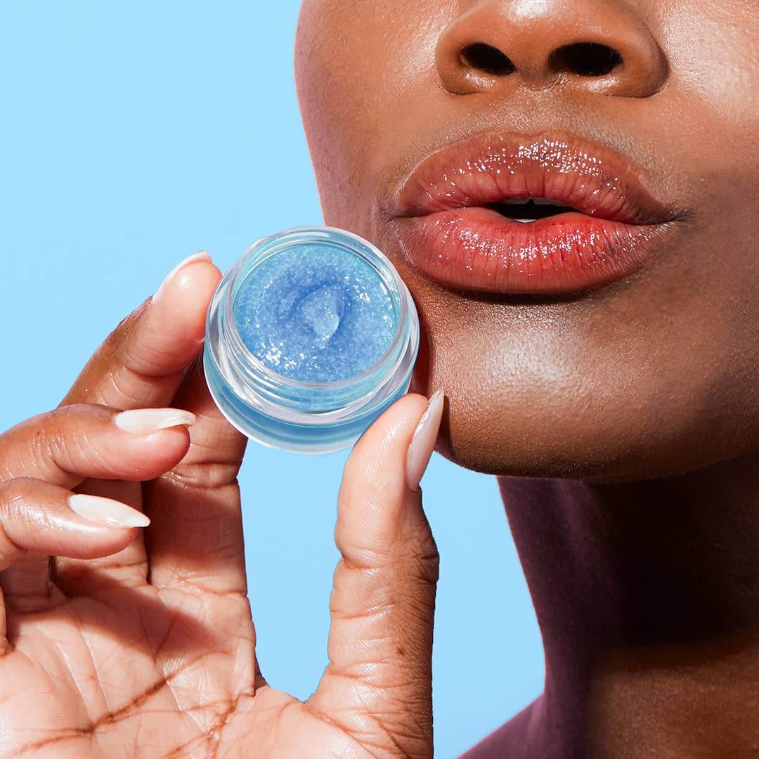 Person holding a small container of blue lip balm close to their lips against a clear background