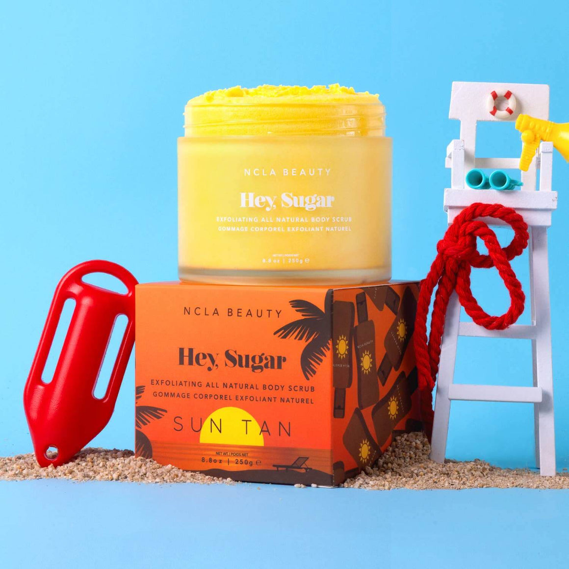 NCLA Beauty 'Hey Sugar' body scrub with packaging on a sandy surface against a blue background