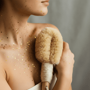 Person holding a dry brush with gold glitter on skin against a neutral background