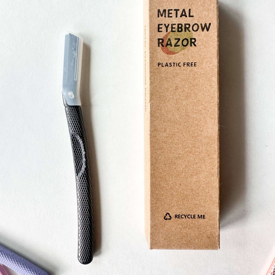 Metal eyebrow razor next to its cardboard box on a white background