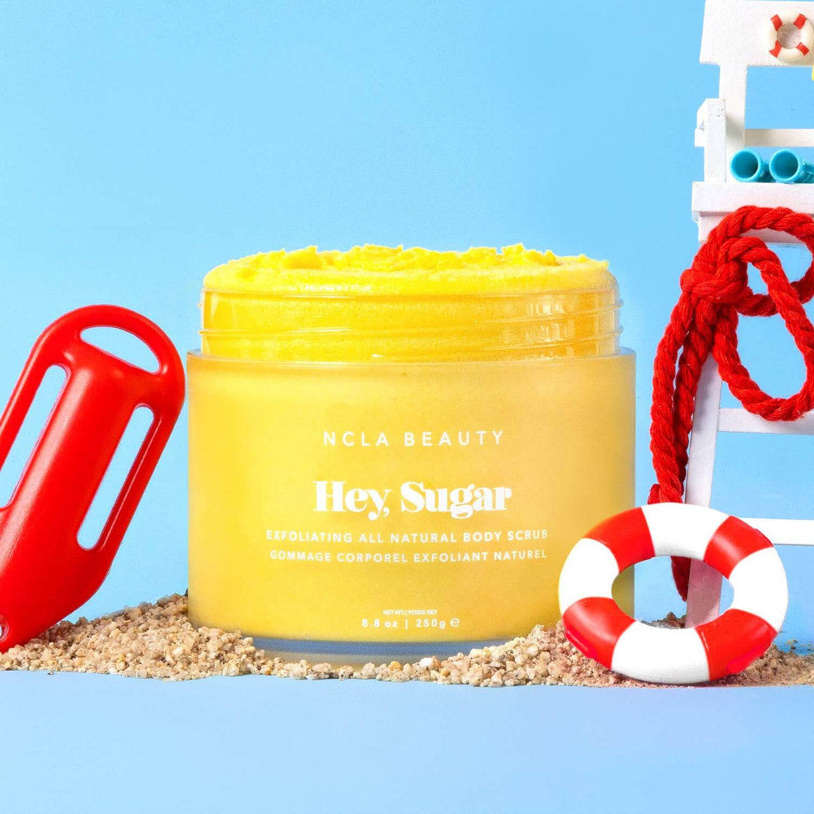 Yellow jar of NCLA Beauty Hey Sugar body scrub with beach-themed props on a blue background