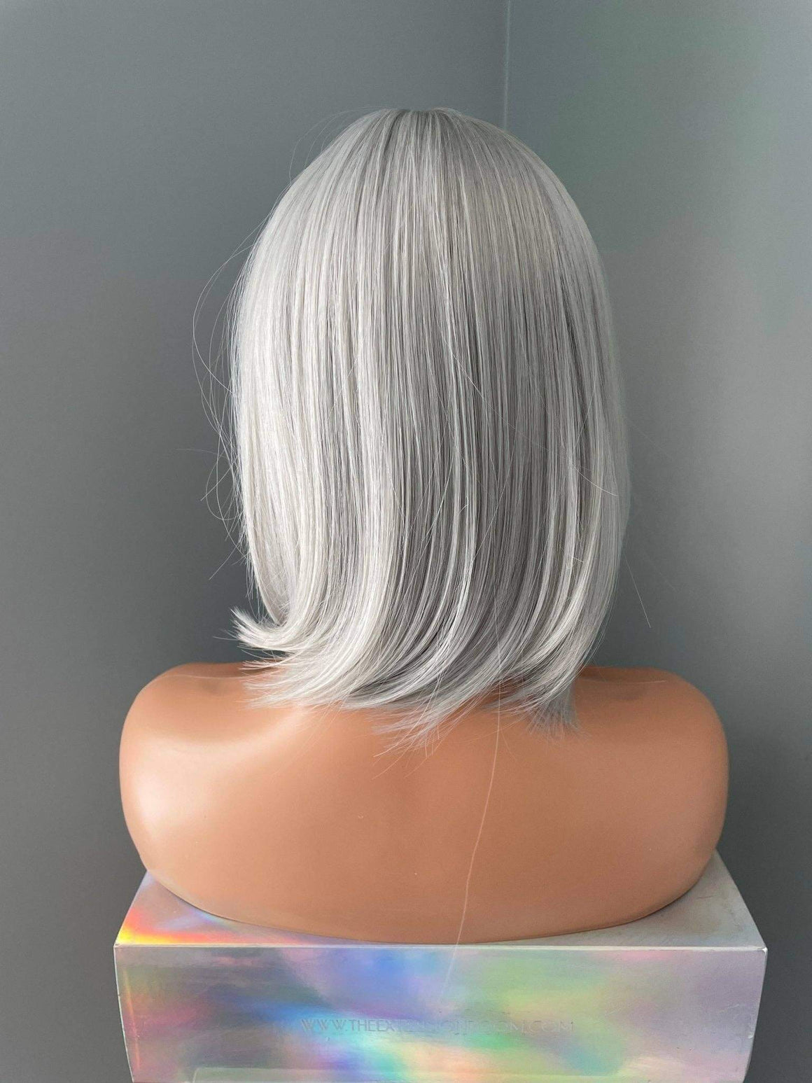 A short, straight wig with silver-gray and white highlights, shown from the back on a mannequin head mounted on a holographic display block against a gray background.